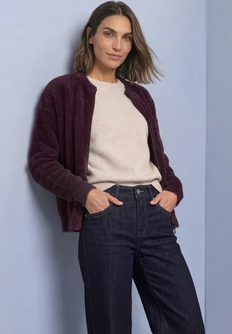 Frau in dunkelrotem Strickcardigan, hellem Pullover und Jeans vor hellem Hintergrund