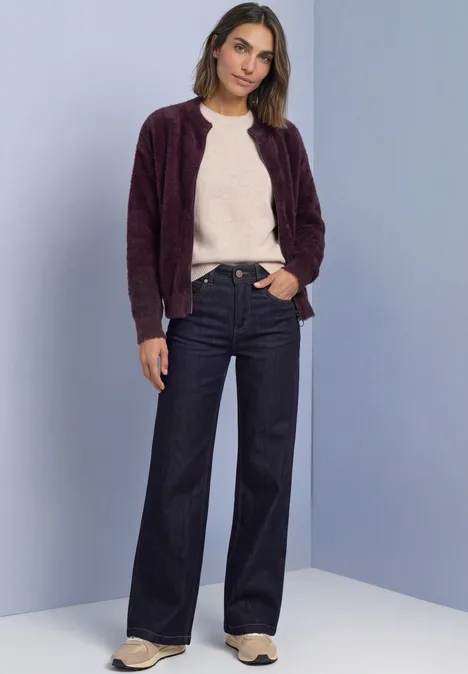 Frau trägt beigefarbenen Pullover, lila Strickjacke und weite Jeans, steht vor neutralem Hintergrund.
