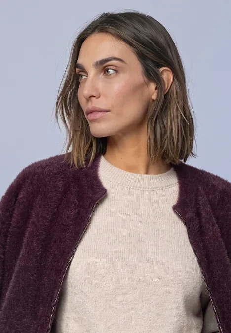 Frau mit kurzen braunen Haaren trägt hellen Pullover und violette Strickjacke, blickt seitlich.