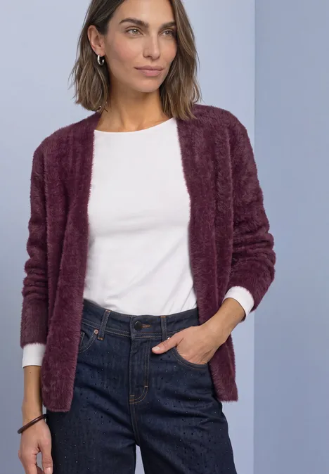 Frau mit weißem Shirt, weinrotem Cardigan und Jeans vor hellem Hintergrund.