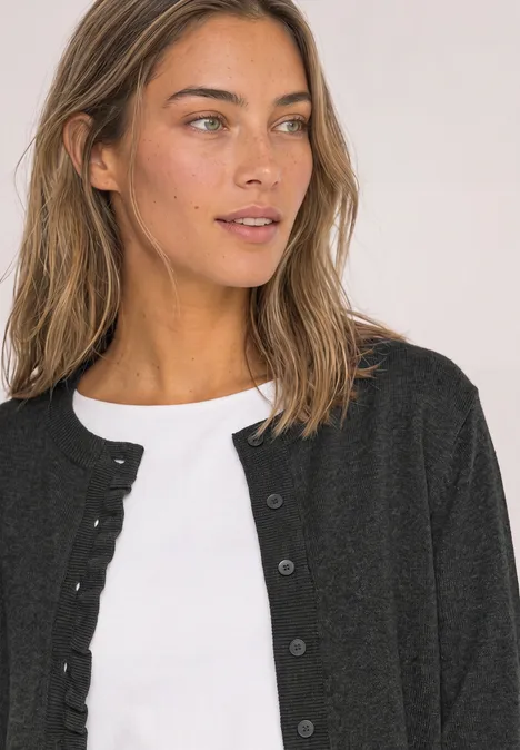 Cardigan à col rond et à volants armor grey mel. Femme aux cheveux bruns portant un top blanc et un cardigan noir, regard de côté.