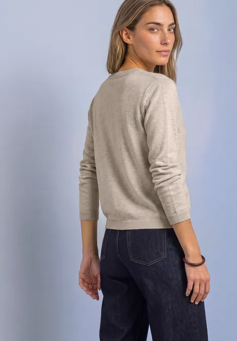 Cardigan à col rond et à volants clay sand mel. Frau in beigem Pullover und dunkler Jeans dreht sich leicht zur Kamera vor hellem Hintergrund.