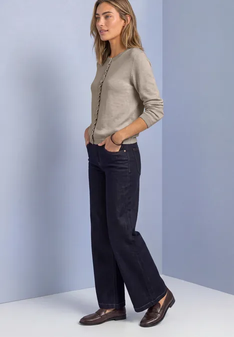 Cardigan à col rond et à volants clay sand mel. Frau in beigem Cardigan, weiten dunklen Hosen und braunen Schuhen vor hellem, zweifarbigem Hintergrund.
