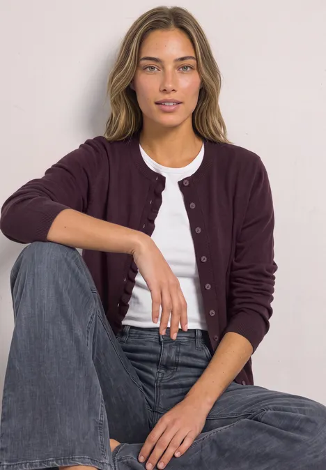 Frau mit bordeauxfarbenem Cardigan, weißem Shirt und Jeans sitzt lässig vor hellem Hintergrund.