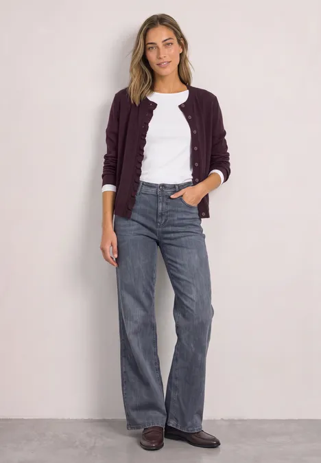Eine Frau steht vor heller Wand, trägt weiße Bluse, bordeauxfarbene Strickjacke, weite graue Jeans und braune Schuhe.