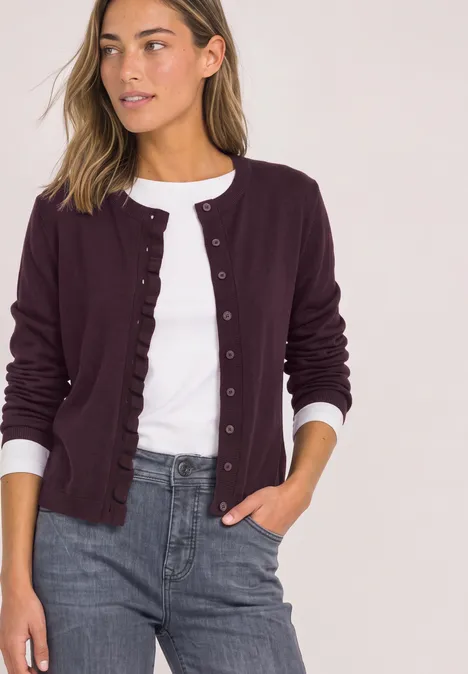 Frau in lila Cardigan mit Rüschen am Saum, weißem Shirt und grauer Hose.
