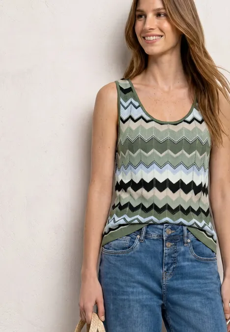 Frau trägt ärmelloses, grün-schwarz-weißes Zickzack-Oberteil und blaue Jeans, hält ein Handy.