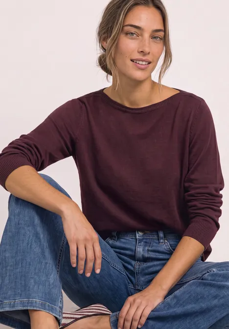Een vrouw zit glimlachend in een bordeaux trui en jeans voor een witte achtergrond.
