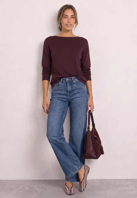 Vrouw in bordeaux trui, blauwe wijde jeans, slippers, bijpassende tas, staand tegen een witte muur.