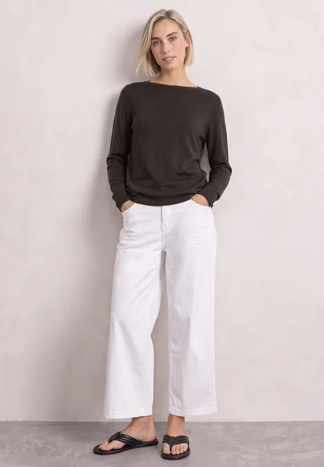 Frau in schwarzem Pullover, weißer weiter Hose und schwarzen Sandalen vor neutralem Hintergrund.