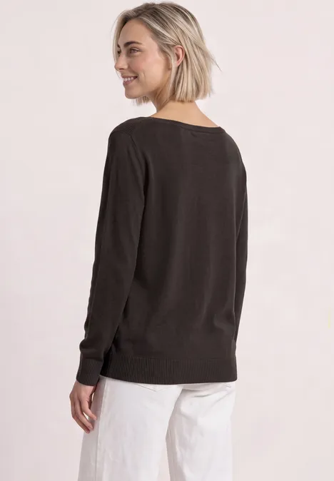 Frau mit kurzem blonden Haar trägt einen dunkelbraunen Pullover und weiße Hose, lächelt und schaut zurück.