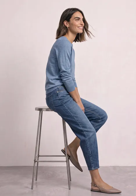 Eenvoudige trui met boothals northern blue mel. Frau sitzt seitlich auf einem Hocker, trägt blauen Pullover, Jeans und flache Schuhe vor hellem Hintergrund.
