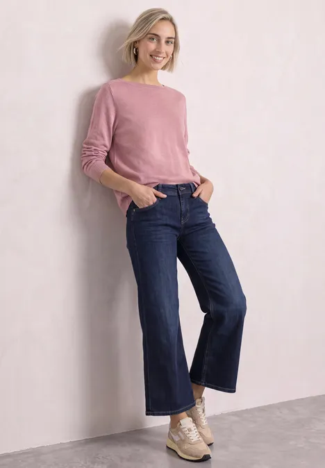 Frau mit rosa Pullover, weitem Jeansmodell und Turnschuhen lehnt an einer Wand und lächelt.