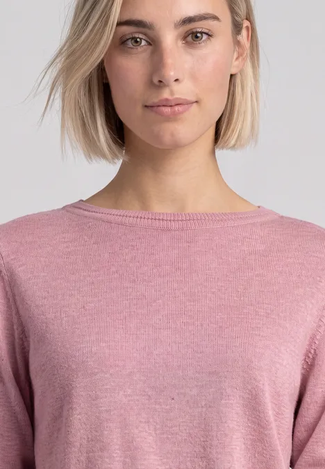 Frau mit kurzen blonden Haaren trägt einen rosafarbenen Pullover vor neutralem Hintergrund.