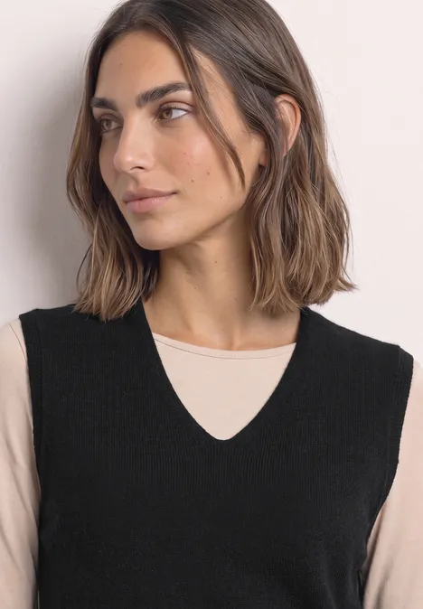 Vrouw in zwart mouwloos vest met V-hals over beige top, zijwaarts kijkend.