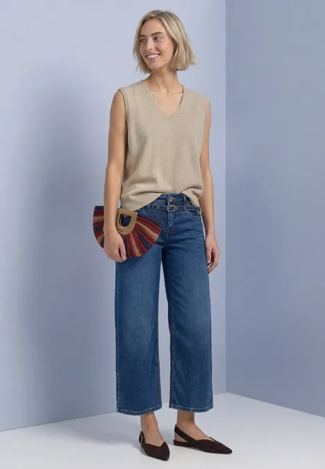 Pullunder mit V-Neck in Unifarbe light sepia sand mel. Frau vor hellviolettem Hintergrund trägt beiges ärmelloses Top, weite Cropped-Jeans, flache Schuhe und runde Tasche.