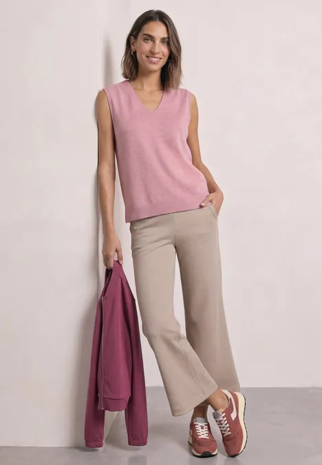 Pullunder mit V-Neck in Unifarbe vintage blush mel. Frau in rosafarbenem ärmellosem Pullover, beigen Schlaghosen und Sneakers, hält eine pinke Jacke.