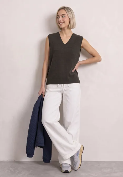 Pullunder mit V-Neck in Unifarbe black coffee mel. Frau mit blondem Bob trägt ärmelloses schwarzes Top, weiße Schlaghose und Sneaker, hält blauen Blazer.