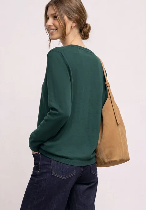 Frau in grünem Langarmshirt, Jeans und beiger Tasche, seitlich stehend, lässiges Styling.