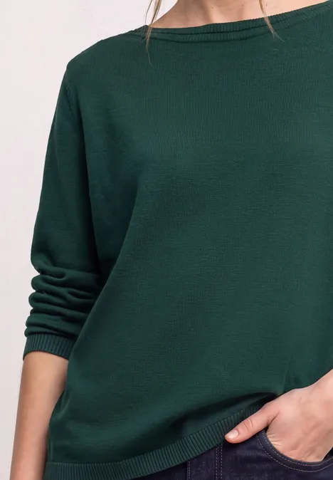 Person trägt locker geschnittenen dunkelgrünen Pullover mit hochgekrempelten Ärmeln, Hand in Tasche.