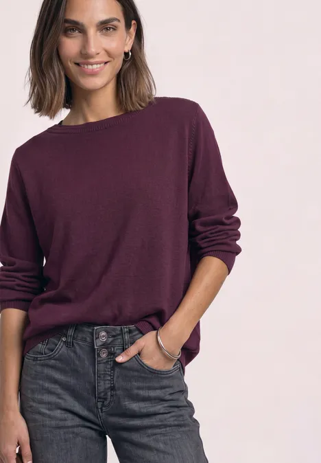 Frau mit lila Pullover und dunklen Jeans vor hellem Hintergrund.