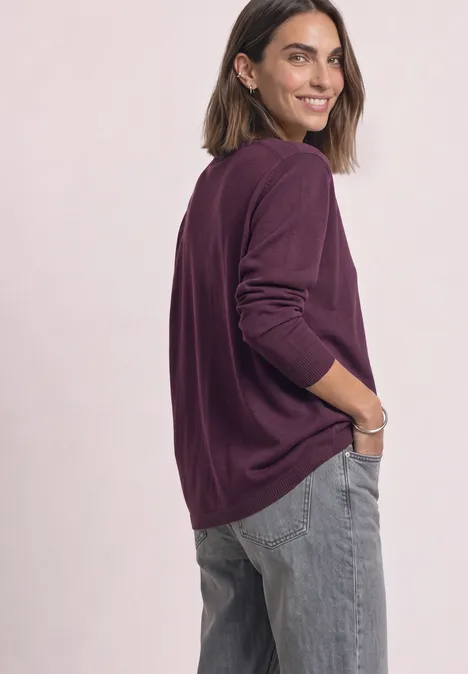 Frau im violetten Pullover und grauen Jeans blickt lächelnd über die Schulter.