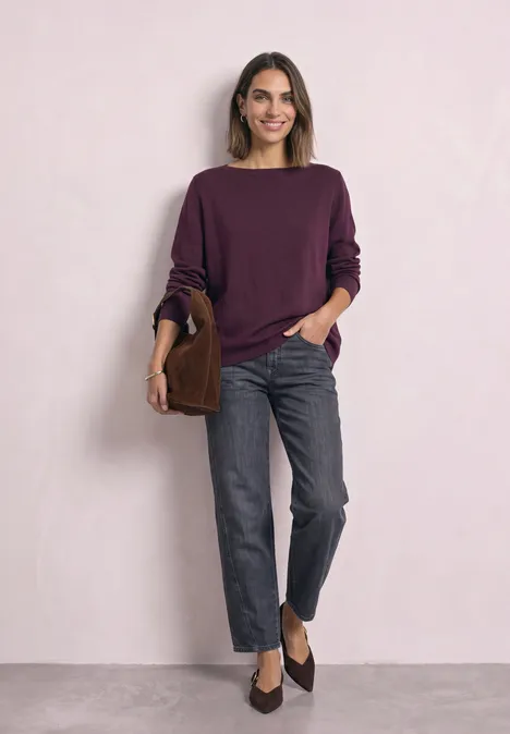 Frau in violettem Pullover, grauen Jeans und dunklen Slippern, mit brauner Tasche vor hellem Hintergrund.