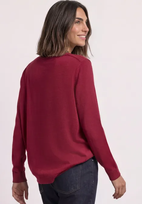 Frau in bordeauxrotem Pullover und Jeans, lächelt über die Schulter.