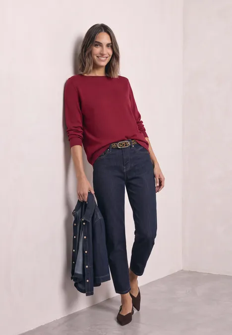 Frau lehnt lächelnd an heller Wand, trägt roten Pullover, dunkle Jeans, Gürtel und hält Jeansjacke