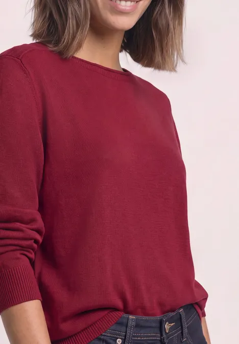 Frau trägt einen roten Pullover, halbe Körperansicht vor hellem Hintergrund.
