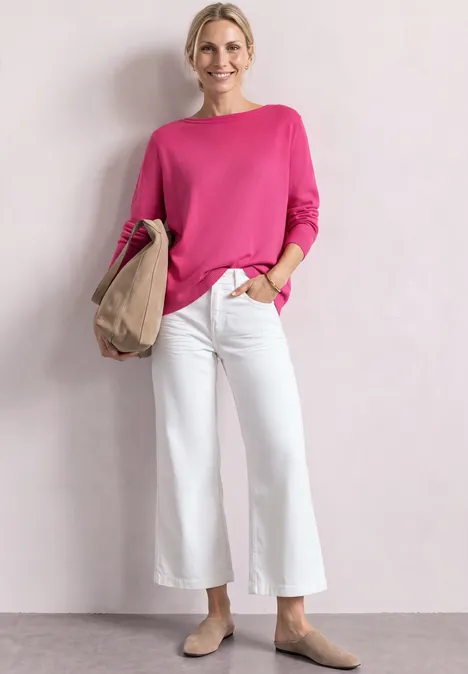 Frau in pinkem Pullover, weißer Culotte-Jeans und beigen Schuhen hält Papiertüte und lächelt.