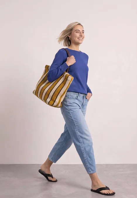 Frau in blauer Bluse und Jeans, mit gestreifter Tasche und Sandalen, geht glücklich spazieren.