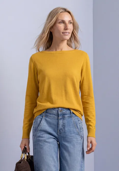 Frau in senfgelbem Pullover und blauen Jeans hält eine Tasche und schaut zur Seite.