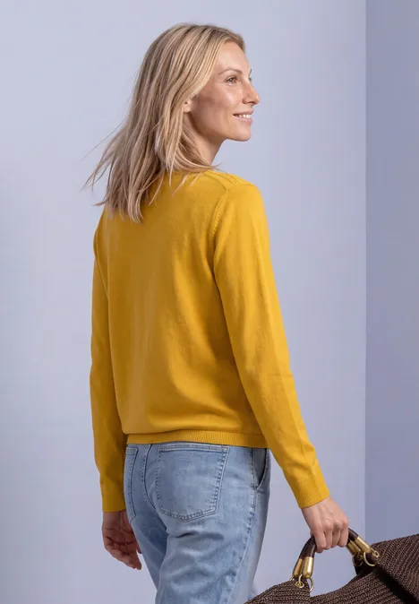 Frau in gelbem Pullover und Jeans hält lächelnd einen braunen Korb in der Hand.