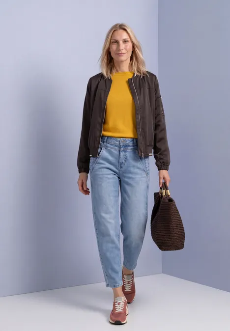 Frau in gelbem Shirt, brauner Jacke, hellblauen Jeans und Sneakers hält eine braune Tasche vor hellem Hintergrund.