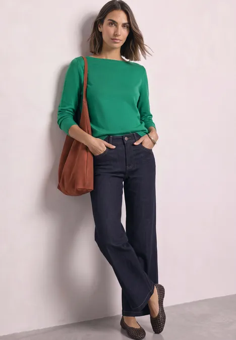 Frau mit grünem Langarmshirt, dunklen Jeans, brauner Umhängetasche und Loafers lehnt an einer Wand.