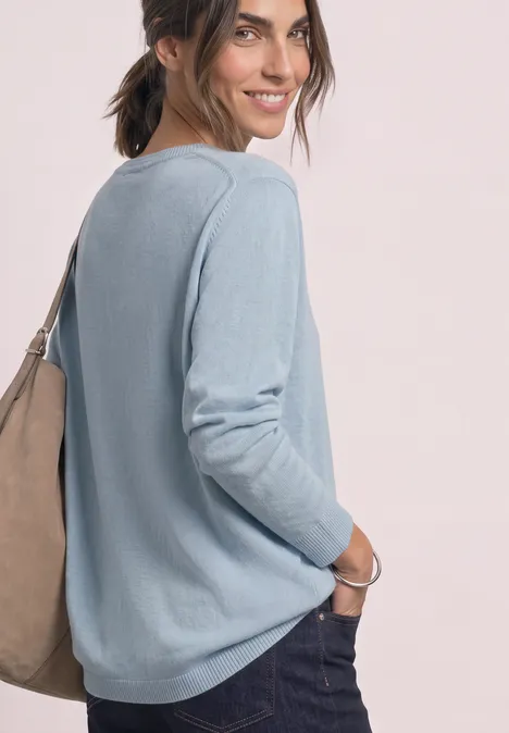 Frau im hellblauen Langarmshirt mit lässiger Jeans und Umhängetasche, lächelnd seitlich stehend.