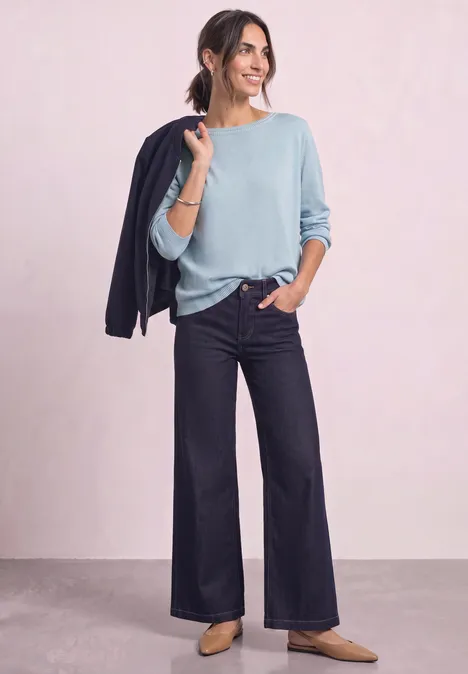 Frau in hellblauem Pullover und weitem Jeans, lässig, mit dunkelblauer Jacke über der Schulter.