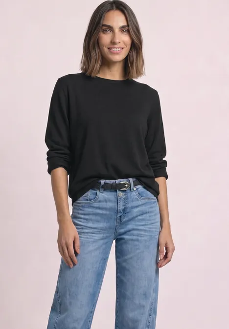 Eine Frau mit schwarzem Pullover und blauer Jeans posiert vor hellem Hintergrund.