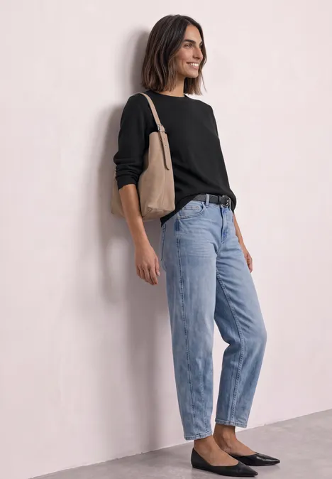 Frau lehnt an Wand, trägt schwarzen Pullover, hellblaue Jeans, schwarze Ballerinas und beige Tasche.