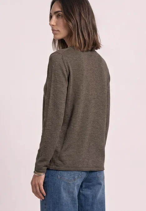 Frau in braunem Langarmshirt und blauen Jeans, steht seitlich vor hellem Hintergrund und blickt zur Seite.