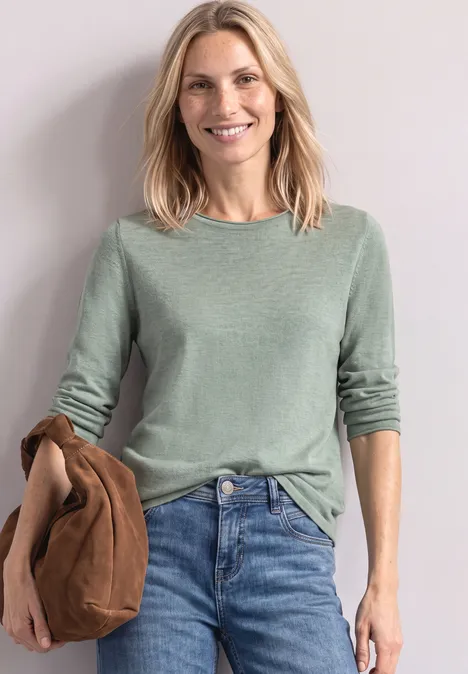 Eine lächelnde, blonde Frau in grünem Pullover und Jeans, die eine braune Jacke hält.