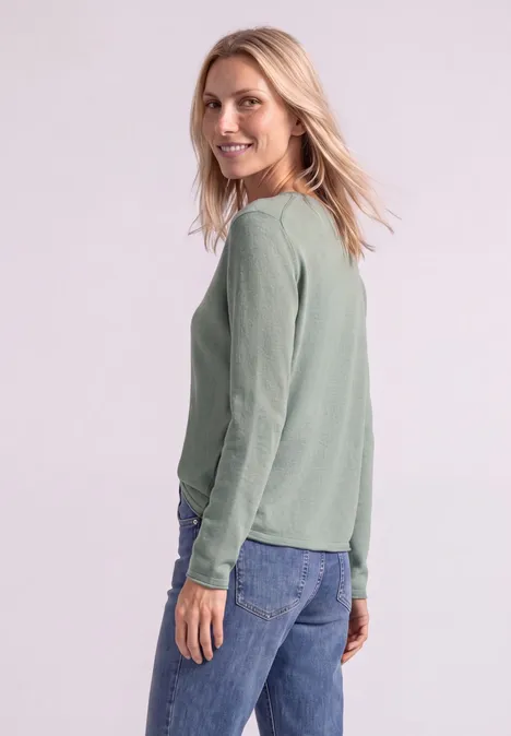 Seitlich fotografierte Frau mit langem, grünem Pullover und Jeans, die lächelnd über die Schulter blickt.
