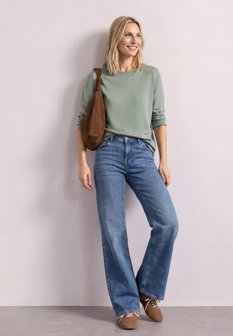 Eine lächelnde Frau in grünem Pullover, weitem, hochgeschnittenem Jeans, trägt braune Tasche und Schuhe.