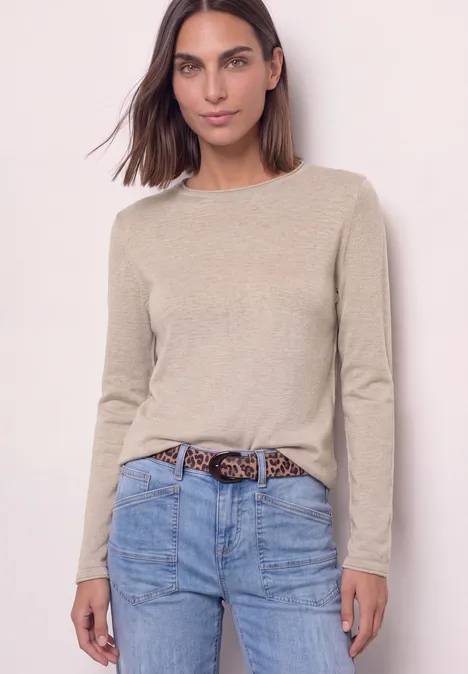 Eine Frau im beigefarbenen Langarmshirt und Jeans mit Leopardenmuster-Gürtel steht vor hellem Hintergrund.