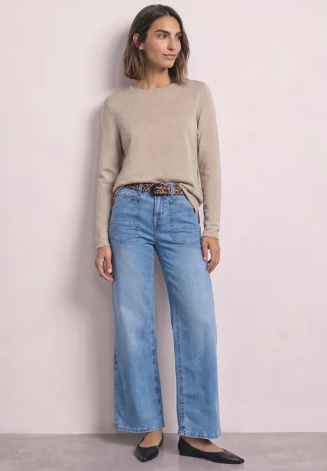 Frau in beigem Pullover, weiten hellblauen Jeans mit braunem Gürtel und schwarzen Schuhen vor hellem Hintergrund.