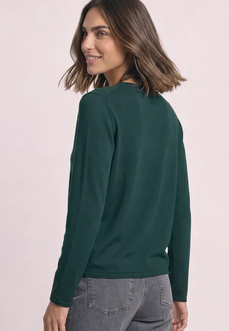 Junge Frau mit schulterlangen braunen Haaren lächelt über die Schulter, trägt einen dunkelgrünen Pullover und graue Jeans.