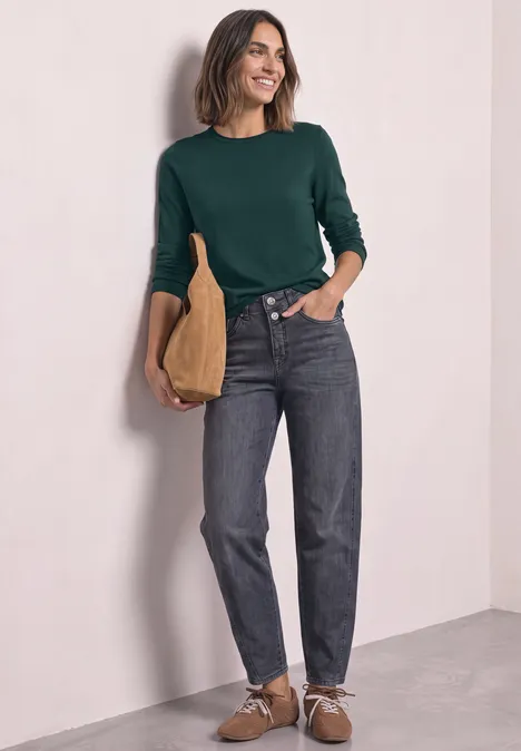 Eine Frau mit grüner Bluse und Jeans lehnt lächelnd an einer Wand und hält eine beige Tasche.