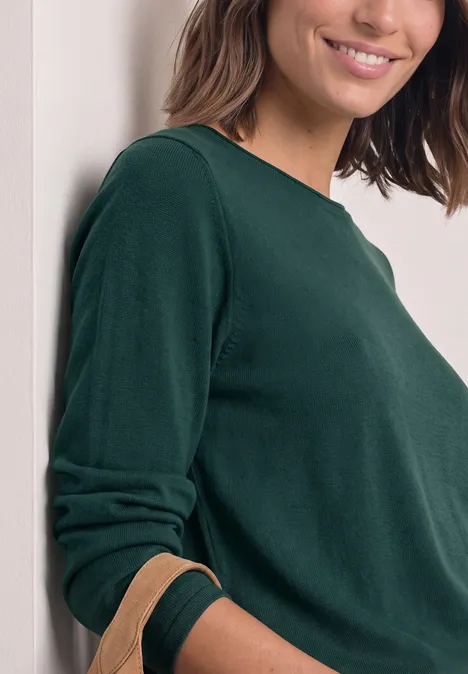 Eine Frau lehnt an einer Wand, trägt ein dunkelgrünes Langarmshirt und lächelt sanft.