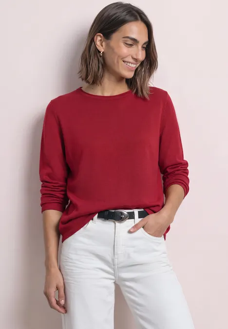 Frau in rotem Langarmshirt und weißen Jeans, lächelnd mit einer Hand in der Tasche.